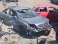 Zniszczony szary Opel, z otwartą klapą bagażnika i pękniętą przednią szybą, czeka na złomowanie. Obok czerwony samochód.