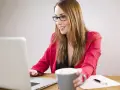 Uśmiechnięta kobieta w okularach pracuje przy laptopie, trzymając kubek. Planuje dokupienie AC do swojego ubezpieczenia.