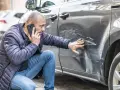Mężczyzna sprawdza telefonem głębokie zarysowania na samochodzie, zgłaszając szkoda komunikacyjna.