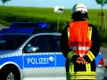 Strażak w kamizelce odblaskowej stoi obok radiowozu z napisem "POLIZEI". Służy jako policjant kierujący ruchem.