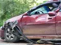 Rozbity czerwony samochód z pękniętą szybą i wgnieceniami. Wygląda na to, że jest to szkoda całkowita, a dalsze użytkowanie pojazdu jest niemożliwe.