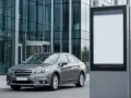 Szary sedan przed nowoczesnym budynkiem. Czy można zarejestrować samochód online? Pusta tablica reklamowa obok.