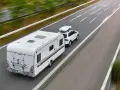 Biały samochód z boxem dachowym ciągnie przyczepę kempingową. To właśnie co nazywamy zespołem pojazdów, jadącym autostradą.