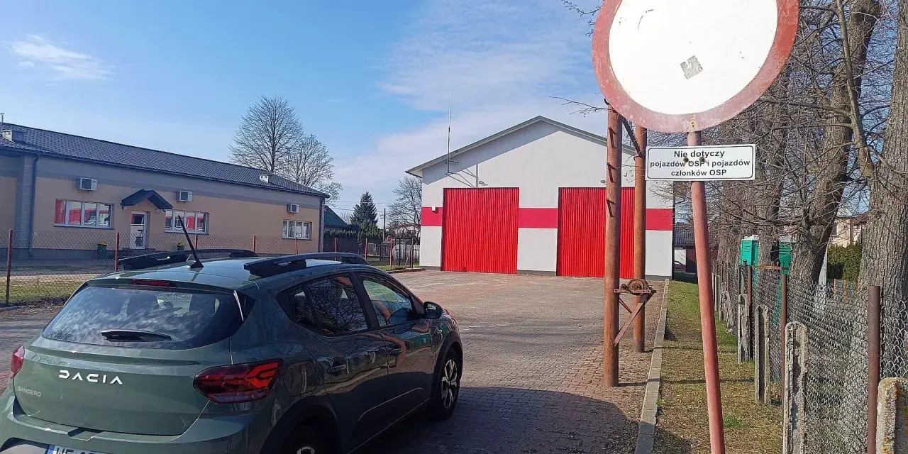 Zielony Dacia Sandero zaparkowany przy remizie OSP. Znak zakazu wjazdu, który może oznaczać mandat za wjazd pod zakaz, jest widoczny obok.