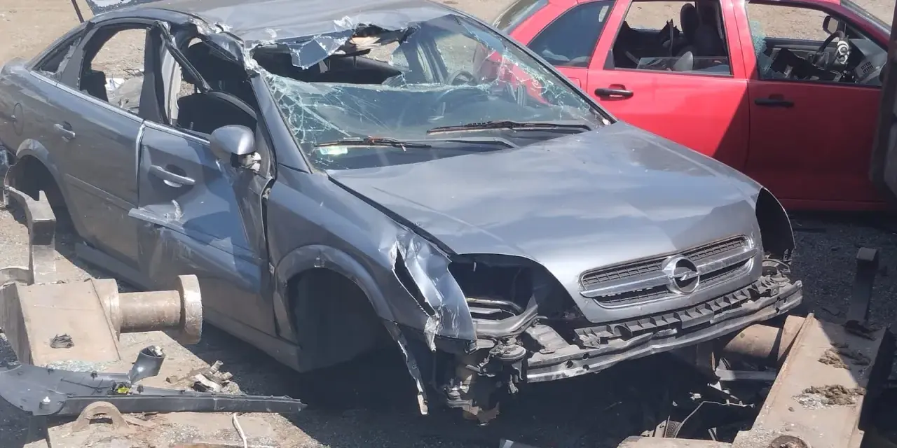Zniszczony szary Opel, z otwartą klapą bagażnika i pękniętą przednią szybą, czeka na złomowanie. Obok czerwony samochód.