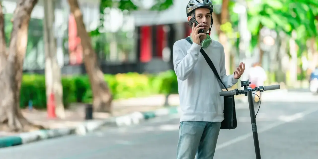 Młody mężczyzna w kasku rozmawia przez telefon, stojąc obok hulajnogi elektrycznej. Czy na hulajnodze elektrycznej trzeba mieć kask? On ma, więc chyba tak!