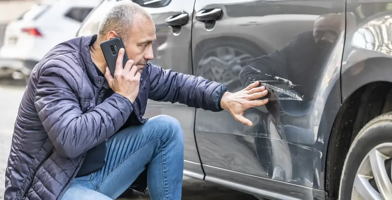 Mężczyzna sprawdza telefonem głębokie zarysowania na samochodzie, zgłaszając szkoda komunikacyjna.