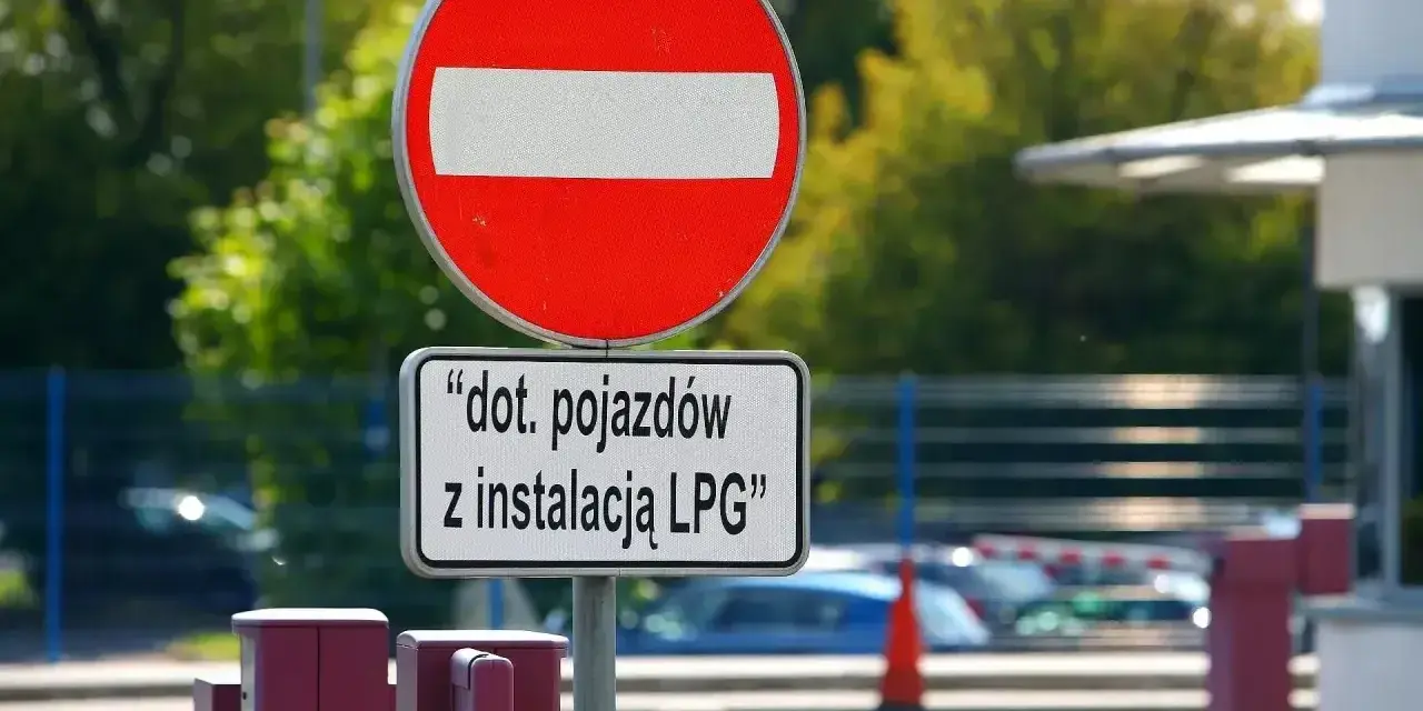 Zakaz wjazdu znak dla pojazdów z instalacją LPG. Barierka i budynek w tle.