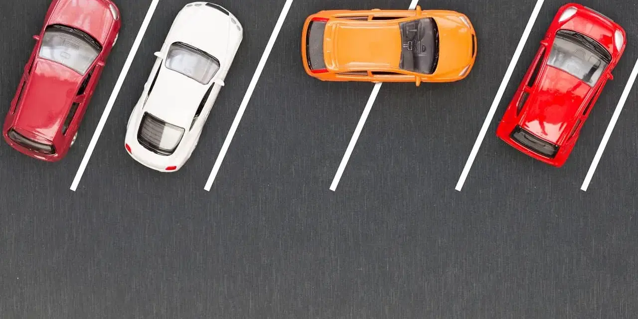 Czerwony, biały i pomarańczowy samochodzik na parkingu. Wykonują parkowanie równoległe przodem, zajmując miejsca wyznaczone białymi liniami.