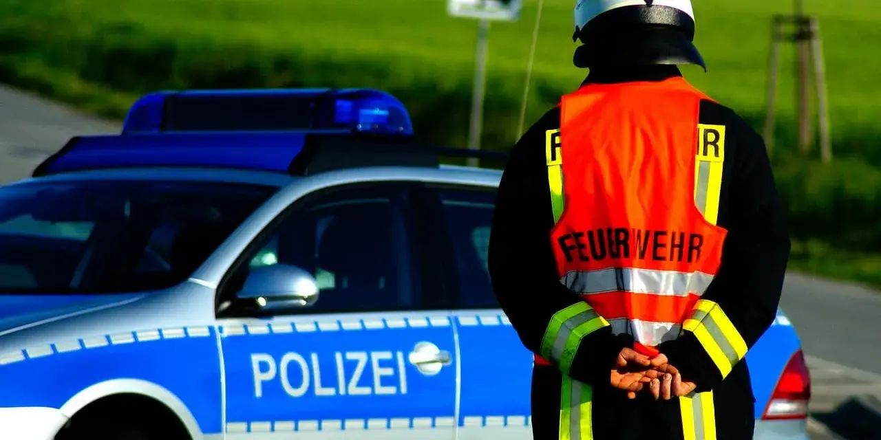 Strażak w kamizelce odblaskowej stoi obok radiowozu z napisem "POLIZEI". Służy jako policjant kierujący ruchem.