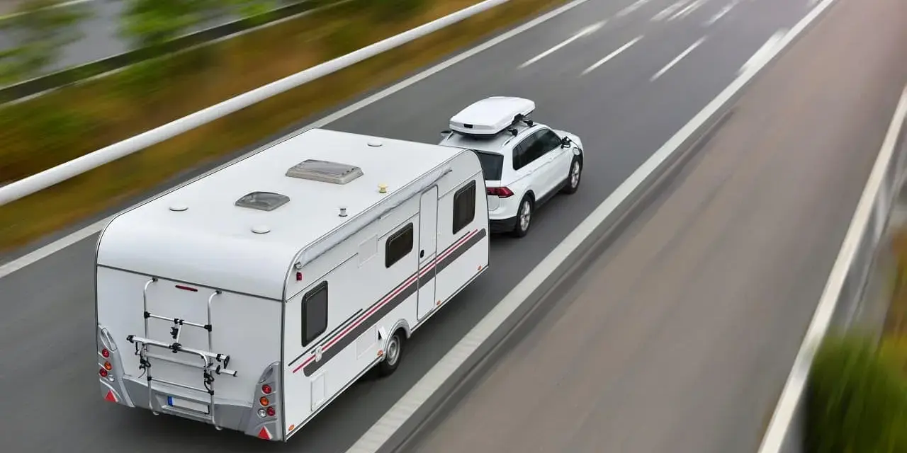 Biały samochód z boxem dachowym ciągnie przyczepę kempingową. To właśnie co nazywamy zespołem pojazdów, jadącym autostradą.