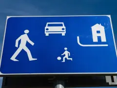 Znak strefa zamieszkania: pieszy, samochód, dziecko grające w piłkę i dom. Bezpieczna przestrzeń dla wszystkich.