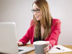 Uśmiechnięta kobieta w okularach pracuje przy laptopie, trzymając kubek. Planuje dokupienie AC do swojego ubezpieczenia.