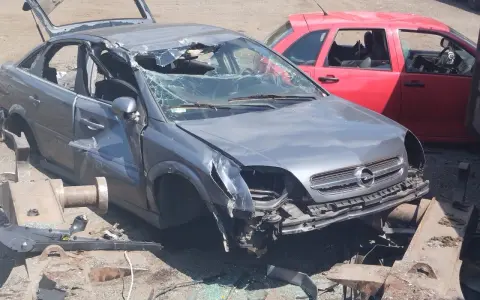 Zniszczony szary Opel, z otwartą klapą bagażnika i pękniętą przednią szybą, czeka na złomowanie. Obok czerwony samochód.
