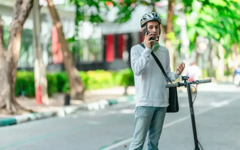 Młody mężczyzna w kasku rozmawia przez telefon, stojąc obok hulajnogi elektrycznej. Czy na hulajnodze elektrycznej trzeba mieć kask? On ma, więc chyba tak!