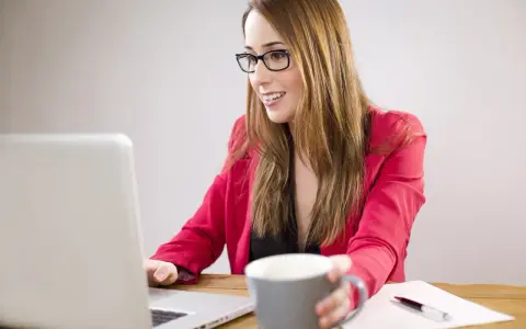 Uśmiechnięta kobieta w okularach pracuje przy laptopie, trzymając kubek. Planuje dokupienie AC do swojego ubezpieczenia.