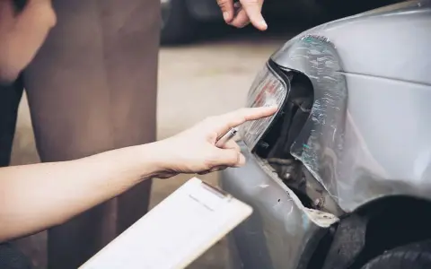 Dwóch mężczyzn ocenia szkody parkingowe na samochodzie. Jeden wskazuje palcem na uszkodzony reflektor, drugi trzyma dokumenty.