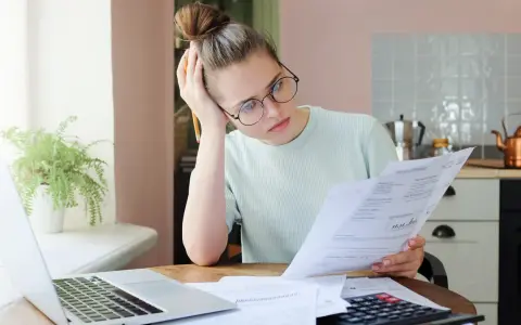 Zmartwiona kobieta analizuje dokumenty, szukając informacji o zwrocie OC po sprzedaży samochodu. Obok laptop i kalkulator.