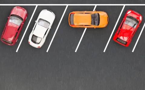 Czerwony, biały i pomarańczowy samochodzik na parkingu. Wykonują parkowanie równoległe przodem, zajmując miejsca wyznaczone białymi liniami.