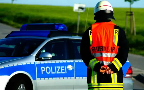 Strażak w kamizelce odblaskowej stoi obok radiowozu z napisem "POLIZEI". Służy jako policjant kierujący ruchem.