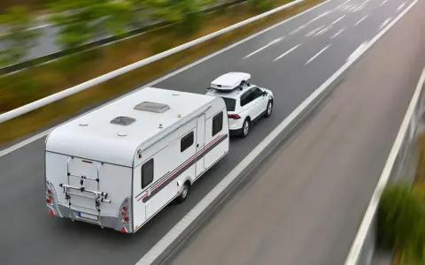 Biały samochód z boxem dachowym ciągnie przyczepę kempingową. To właśnie co nazywamy zespołem pojazdów, jadącym autostradą.