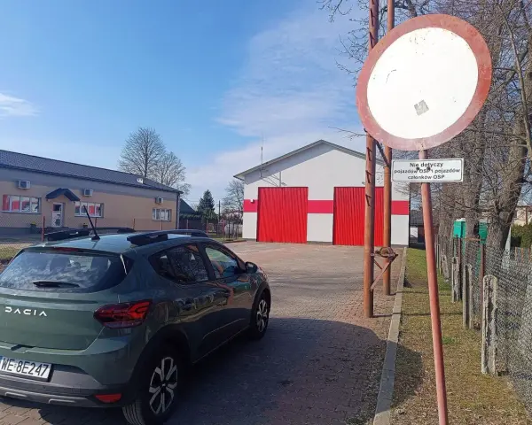 Zielony Dacia Sandero zaparkowany przy remizie OSP. Znak zakazu wjazdu, który może oznaczać mandat za wjazd pod zakaz, jest widoczny obok.