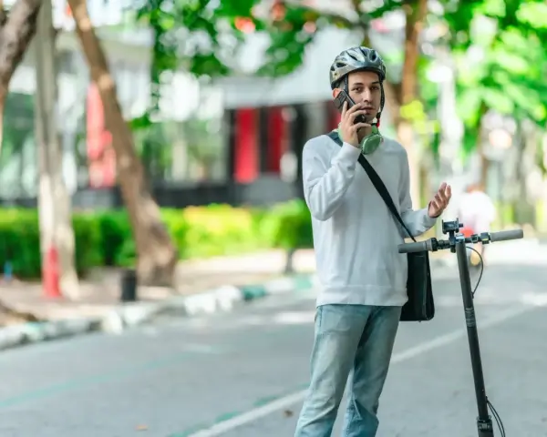 Młody mężczyzna w kasku rozmawia przez telefon, stojąc obok hulajnogi elektrycznej. Czy na hulajnodze elektrycznej trzeba mieć kask? On ma, więc chyba tak!