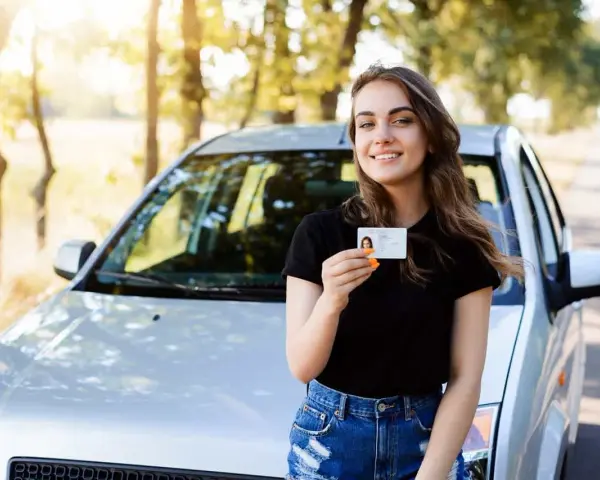 Szczęśliwa młoda kobieta z prawem jazdy 16 lat stoi obok samochodu.