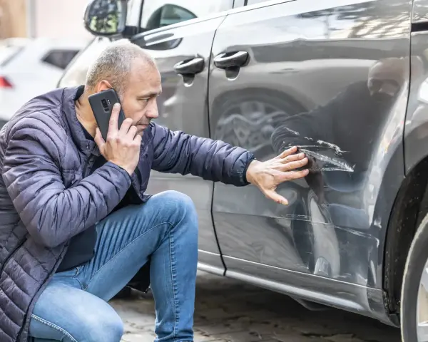 Mężczyzna sprawdza telefonem głębokie zarysowania na samochodzie, zgłaszając szkoda komunikacyjna.