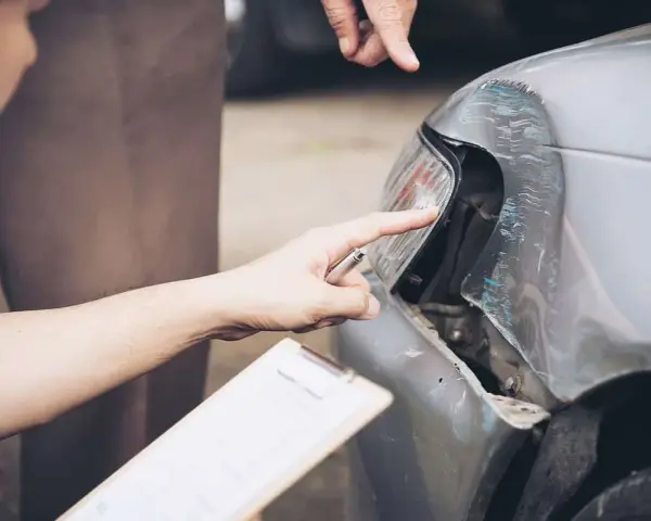 Dwóch mężczyzn ocenia szkody parkingowe na samochodzie. Jeden wskazuje palcem na uszkodzony reflektor, drugi trzyma dokumenty.