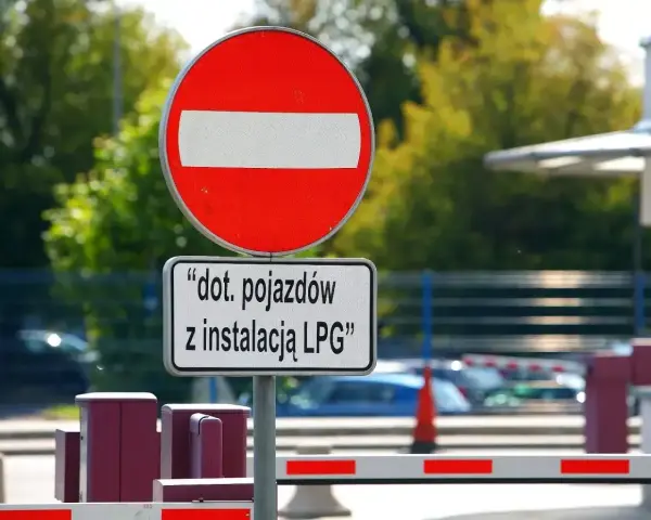 Zakaz wjazdu znak dla pojazdów z instalacją LPG. Barierka i budynek w tle.