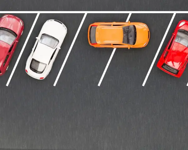 Czerwony, biały i pomarańczowy samochodzik na parkingu. Wykonują parkowanie równoległe przodem, zajmując miejsca wyznaczone białymi liniami.
