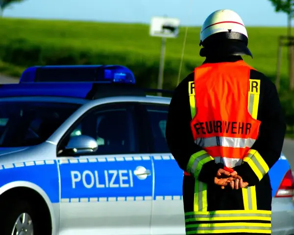 Strażak w kamizelce odblaskowej stoi obok radiowozu z napisem "POLIZEI". Służy jako policjant kierujący ruchem.