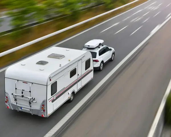 Biały samochód z boxem dachowym ciągnie przyczepę kempingową. To właśnie co nazywamy zespołem pojazdów, jadącym autostradą.