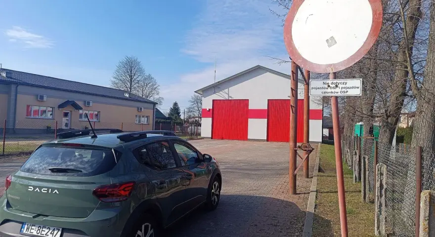 Zielony Dacia Sandero zaparkowany przy remizie OSP. Znak zakazu wjazdu, który może oznaczać mandat za wjazd pod zakaz, jest widoczny obok.