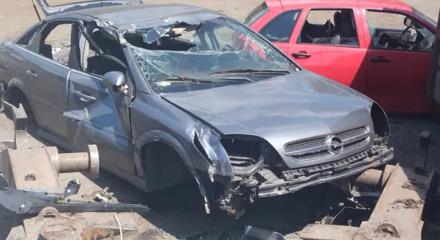 Zniszczony szary Opel, z otwartą klapą bagażnika i pękniętą przednią szybą, czeka na złomowanie. Obok czerwony samochód.