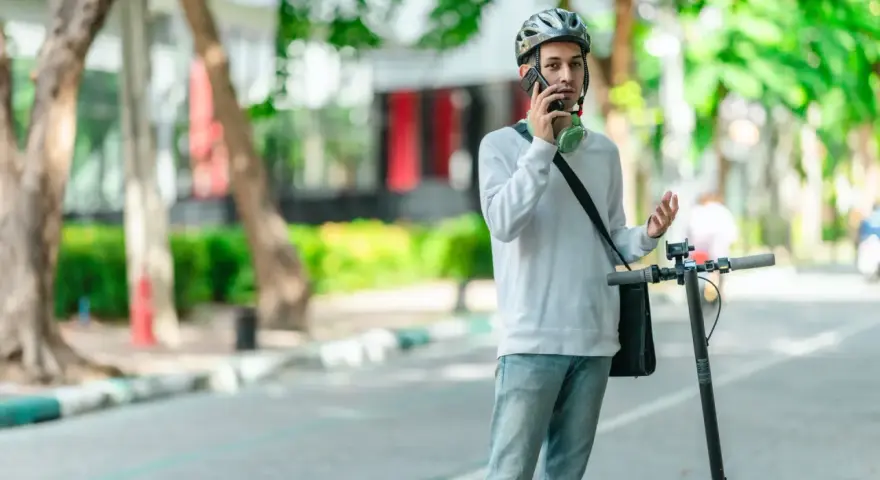 Młody mężczyzna w kasku rozmawia przez telefon, stojąc obok hulajnogi elektrycznej. Czy na hulajnodze elektrycznej trzeba mieć kask? On ma, więc chyba tak!