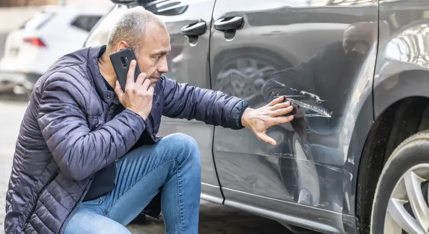 Mężczyzna sprawdza telefonem głębokie zarysowania na samochodzie, zgłaszając szkoda komunikacyjna.