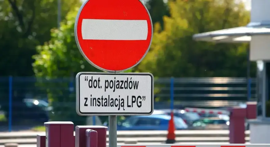 Zakaz wjazdu znak dla pojazdów z instalacją LPG. Barierka i budynek w tle.