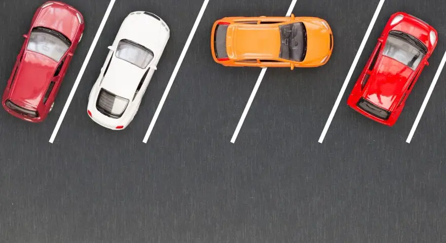 Czerwony, biały i pomarańczowy samochodzik na parkingu. Wykonują parkowanie równoległe przodem, zajmując miejsca wyznaczone białymi liniami.