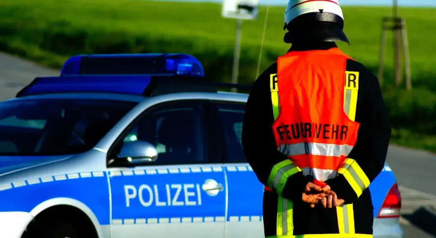 Strażak w kamizelce odblaskowej stoi obok radiowozu z napisem "POLIZEI". Służy jako policjant kierujący ruchem.