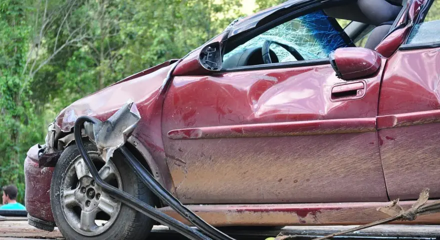 Rozbity czerwony samochód z pękniętą szybą i wgnieceniami. Wygląda na to, że jest to szkoda całkowita, a dalsze użytkowanie pojazdu jest niemożliwe.