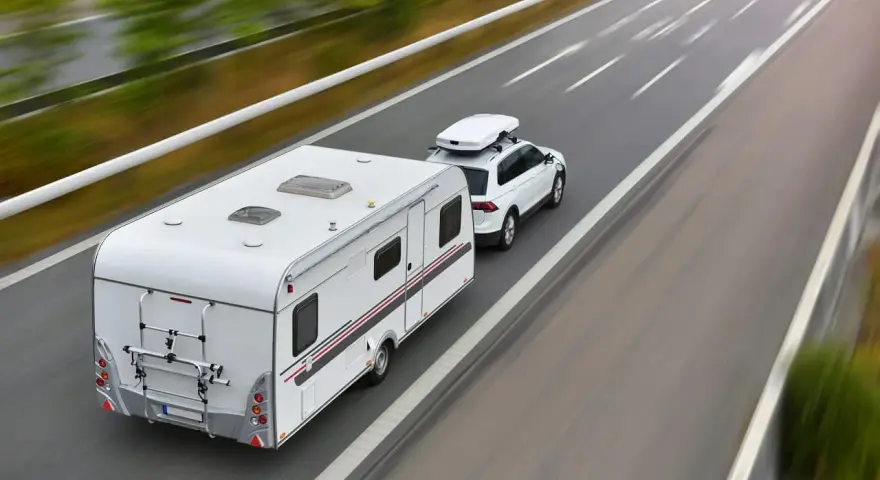 Biały samochód z boxem dachowym ciągnie przyczepę kempingową. To właśnie co nazywamy zespołem pojazdów, jadącym autostradą.