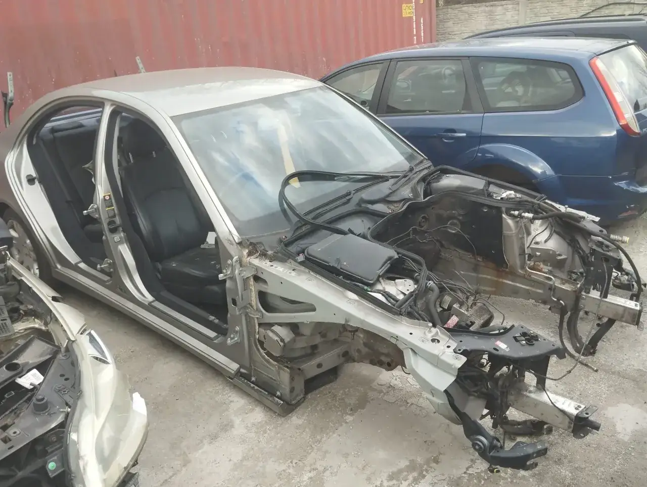 Rozłożony samochód, częściowo rozebrany, jakby ktoś planował jak zezłomować auto. W tle niebieski kombi.