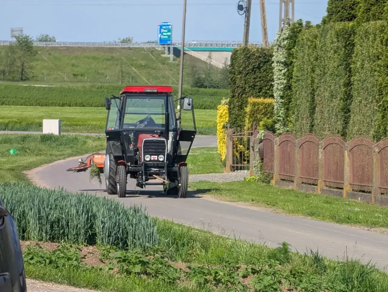 Czerwony traktor z otwartymi drzwiami jedzie po wiejskiej drodze, za nim widać zielone pola i żywopłot.