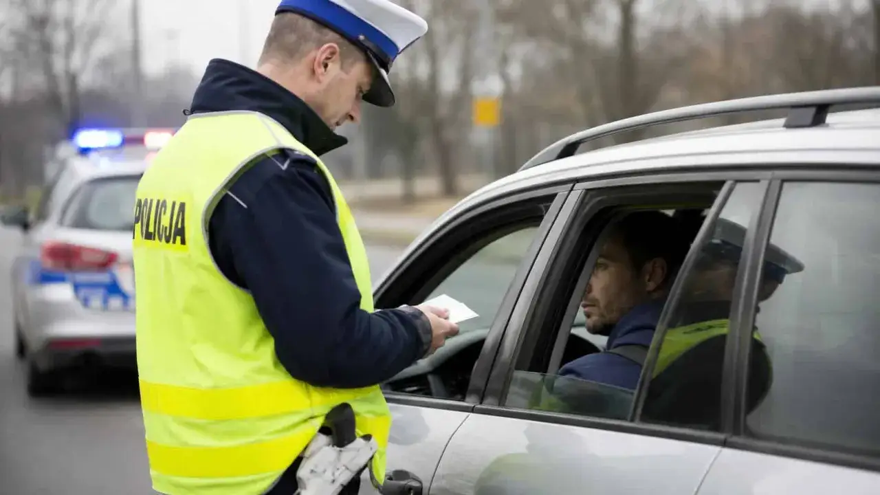 Policjant w kamizelce odblaskowej wręcza dokument kierowcy. W tle radiowóz z włączonymi sygnałami. Może to oznaczać zatrzymanie prawa jazdy na 3 miesiące.