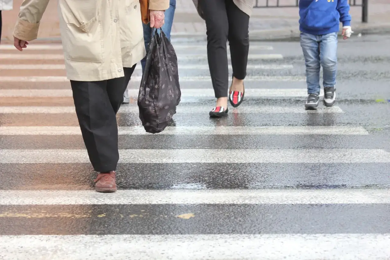 Ludzie przechodzą przez mokre pasy. Starsza pani z torbą, kobieta w czerwono-czarnych balerinkach i dziecko w niebieskiej bluzie. Uważaj na potrącenie na pasach.