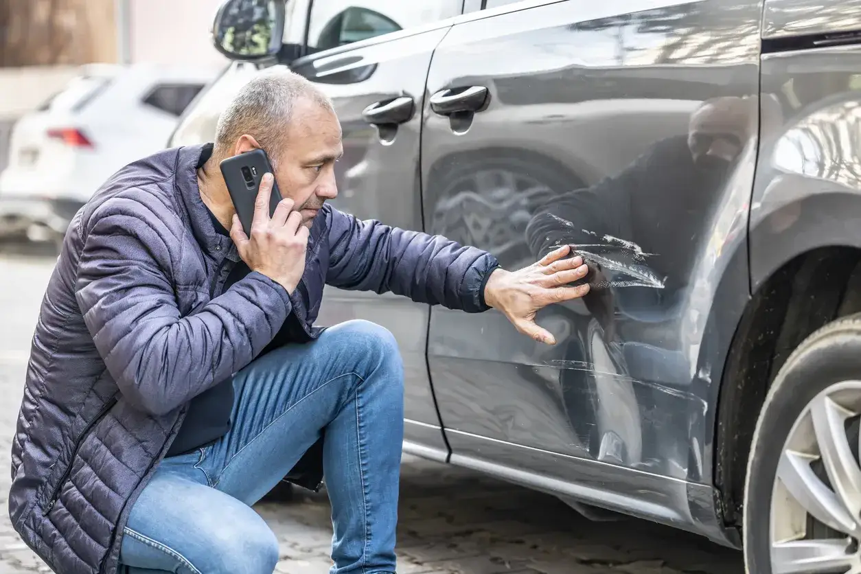 Mężczyzna klęczy przy porysowanym samochodzie, rozmawiając przez telefon. Wygląda na to, że zgłasza szkoda komunikacyjna.