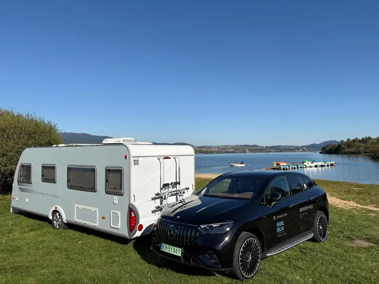 Czarny Mercedes EQE SUV holuje przyczepę kempingową nad jeziorem. B+E co daje? Swobodę podróżowania i relaks na łonie natury.