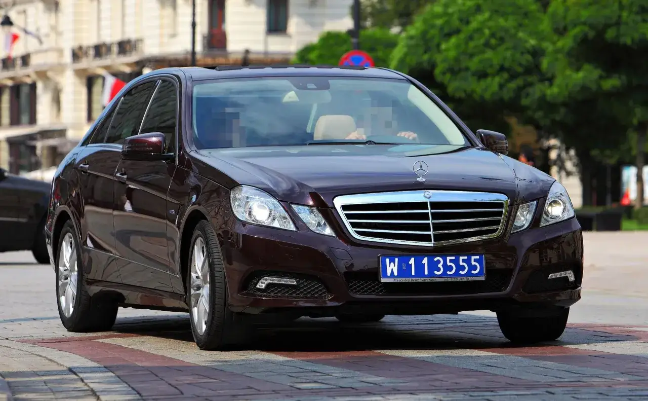 Ciemnobrązowy Mercedes z niebieskimi tablicami rejestracyjnymi W 113555, jadący po ulicy.